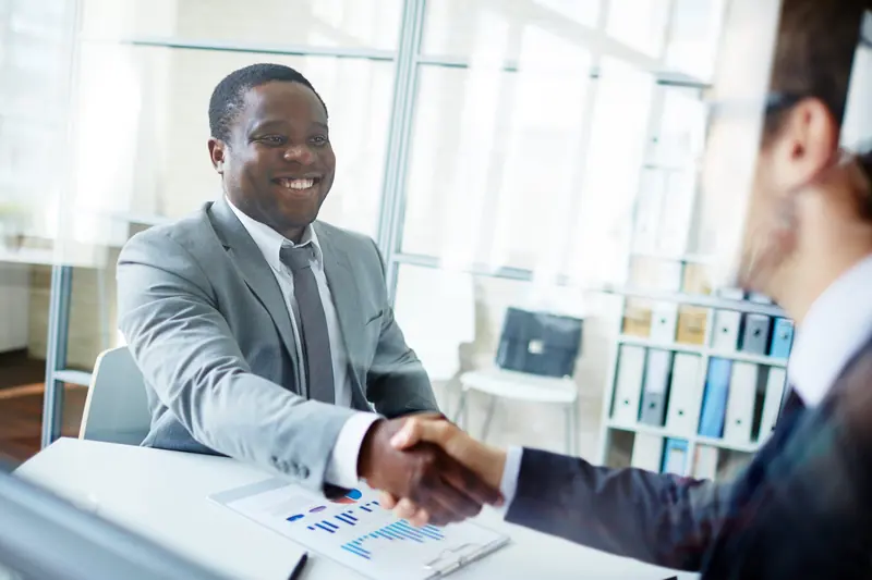 Two men in suits shaking hands across a desk.