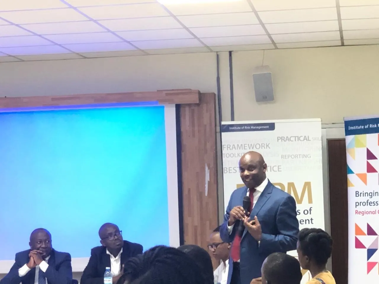 Samuel Kibaara speaking, microphone in hand, beside Institute of Risk Management banners.