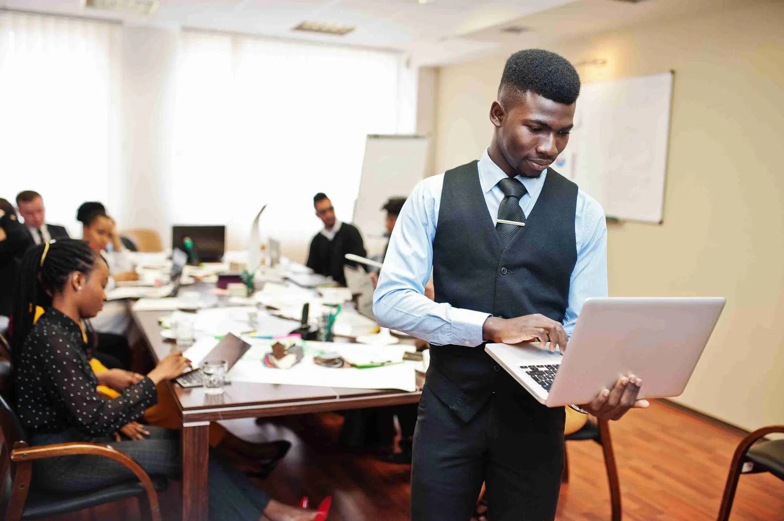 A man standing with a laptop, colleagues at a table behind him.