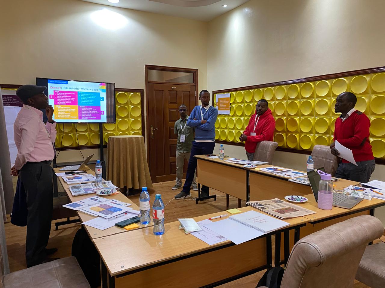 A facilitator stands beside a screen showing an exercise on risk maturity while four delegates stand behind desks; a course workbook lies open in the foreground.